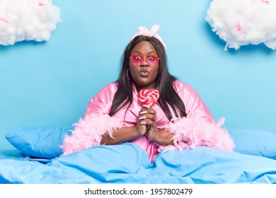 Overweight Dark Skinned Brunette Woman Keeps Lips Folded Holds Delicious Sweet Lollipop Wears Heart Shaped Sunglasses And Dressing Gown Poses On Comfortable Bed Under Warm Blanket Has Lazy Day