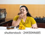 Overweight fat indian woman eating gulab jamun and donuts, laddu or dessert place on a table in kitchen. Unhealthy eating, Craving concept.