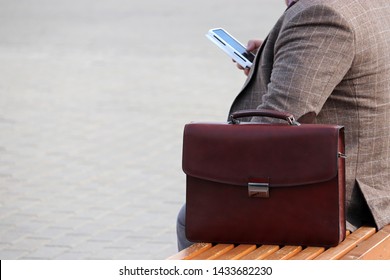 Overweight Man In A Business Suit Sitting With A Leather Briefcase On A Bench With Smartphone In Hand. Concept For Businessman, Official, Government Employee