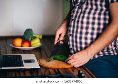 Overweight man looking at recipe using his laptop and cooking in home kitchen. Culinary web blog, weight loss food