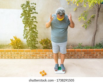 Overweight Man With A Medical Mask Who Weighs Himself On A Scale At Home