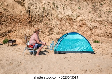 Overweight Man Sits Near Blue Tent On The Beach Or Desert