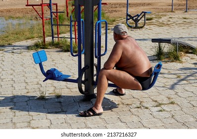 An Overweight Man Sits On A Sports Simulator On The Beach, Does Not Exercise.