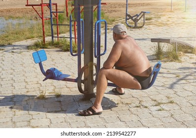 An Overweight Man Is Sitting On A Sports Simulator On The Beach In The Rays Of Light.