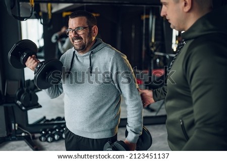 Overweight middle aged men using dumbbells in the gym while training with fitness trainer. Obese man trying to lose weight.