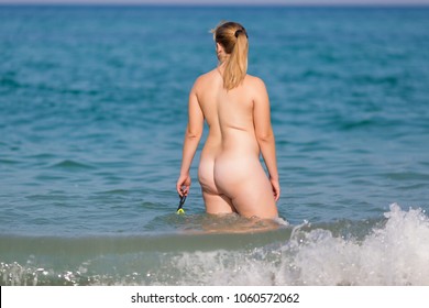 Overweight middle aged woman at the sea. Naked overweight woman enters the sea water with swimming glasses in hand, rear view