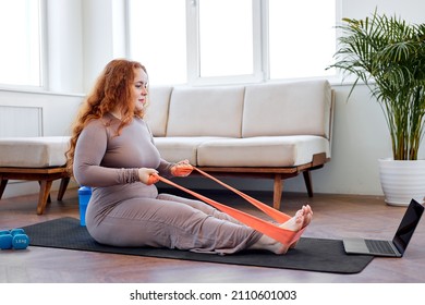 Overweight Redhead Woman Exercising With Rubber Bands According To Online Sports Video On Laptop At Home. Plus Size Caucasian Lady Doing Domestic Workout In Front Of Modern Gadget