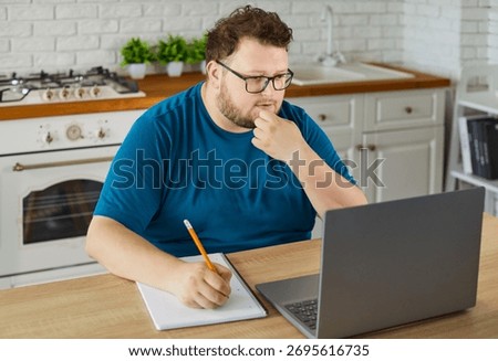 Overweight student using laptop to study and work online at home. Pensive obese person with glasses sitting at kitchen table with notebook and notepad to write notes of remote classes and projects