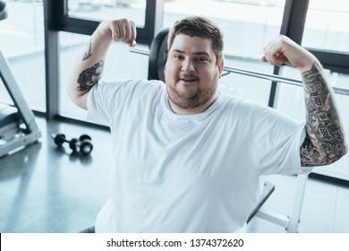 Overweight Tattooed Man Smiling, Looking At Camera And Showing Muscles At Gym