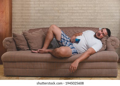 An overweight, unshaven and lazy man of mixed race sleeps on the couch, sprawled out and still holding his cellphone.