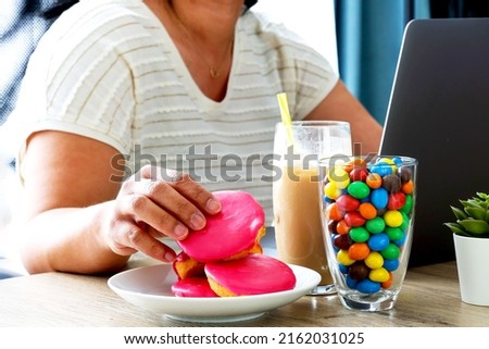 Overweight woman addicted to sweet eating sweet cake, candy hit calories drink while working sitting with laptop at work. 