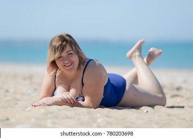 Overweight woman in blue one-piece swimsuit at the sea. Fat girl in blue swimwear lying on sand and looking at camera smiling