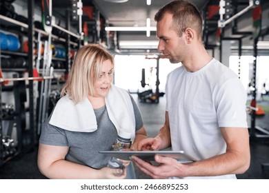 Overweight woman consulting with trainer, discussing daily workout plan. Personal trainer talking with client, discussing exercise program in gym. - Powered by Shutterstock