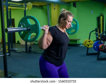 Overweight woman crouches with a barbell in the gym. A fat girl does exercises on the buttocks. Goes in for sports for weight loss.