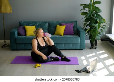 An Overweight Woman Drinks Water From A Bottle Sitting On The Floor On A Mat During An Online Home Sports Workout. Curvy Young Female Doing Pilates For Weight Loss