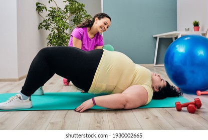 Overweight Woman Exercising Lying On A Yoga Mat, Personal Trainer Assisted While Workout