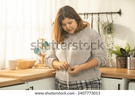 Overweight woman measuring waist with tape on in kitchen with smile of progress for weight lose.