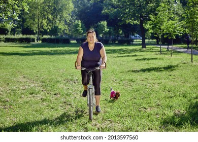 Overweight Woman Is Riding Bicycle While Dog Walking In Public Park In Summer.