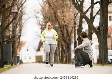 Overweight woman running in park, personal trainer checking her performance. Exercising outdoors for people with obesity, support from friend, fitness coach. - Powered by Shutterstock