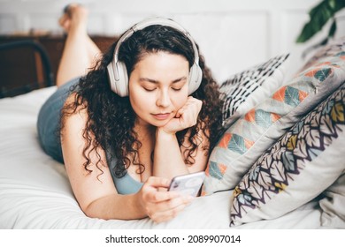 Overweight young woman with loose hair in top and shorts listens to music with wireless headphones holding smartphone sitting on double bed - Powered by Shutterstock