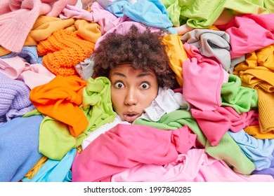 Overwhelmed Afro American woman gives advice to recycle your old clothes sticks out head through multicolored clothing surrouunded by unwearable items collected for donation. Textile recycling
