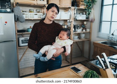 Overwhelmed Asian First Time Mom Is Looking Into The Distance With A Frown While Holding Her Upset Crying Baby Daughter In Arm At A Home Kitchen.