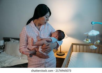 Overwhelmed Asian New Mom Standing By The Baby Bed Is Patting Her Crying Newborn Girl While Trying To Calm Her Down At Midnight In The Bedroom.