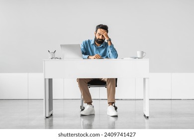 Overwhelmed indian male professional experiencing stress, hand on forehead, while sitting at white desk with laptop in minimalist office, full length - Powered by Shutterstock