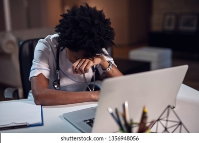 Overworked African American Female Doctor Feeling Tired After Late Night Shift While Sitting At Her Desk And Using Computer. 