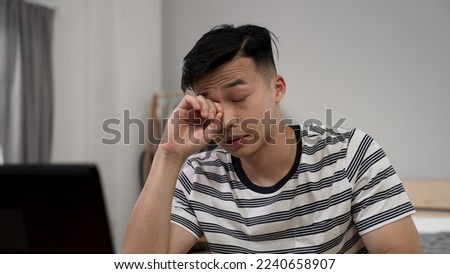 overworked asian college boy learning from home on the computer and rubbing his sore eyes after online class dismissed at desk.