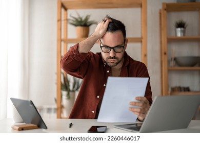 Overworked businessman at home office looking at paper document holding hand on his head feeling hopeless trying to find solution for given problem working too hard need break for better concentration