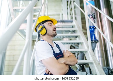 Overworked Caucasian Worker In Overall, With Hardhat And Antiphons Leaning On Railing With Legs Crossed And Sitting On Stairs While Sleeping And Snoring. Night Shift In Factory.