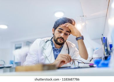 Overworked Doctor In His Office. Tired Male Scientist Trying To Focus, Rubbing His Forehead With Fingers. Mid Adult Male Doctor Working Long Hours. Stressed Male Doctor Sat At His Desk 