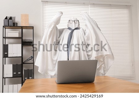 Overworked ghost. Man covered with white sheet using laptop at wooden table in office