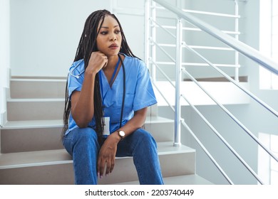 Overworked Nurse In Scrubs Takes Coffee Break In Busy Hospital. Female Nurse Sitting On The Floor And Looking Distraught. Shot Of A Young Nurse Looking Stressed Out While Sitting At A Window