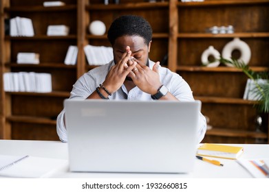 Overworked From Online Work On A Laptop African-American Man Feeling Eye Strain, Burnout, Tired Biracial Male Freelancer Massaging Bridge Of The Nose Sitting At The Workplace In A Modern Office Space