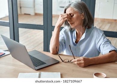 Overworked tired older lady holding glasses feeling headache, having eyesight problem after computer work. Stressed mature senior business woman suffering from fatigue rubbing dry eyes at workplace.