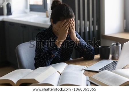 Overworked. Tired young indian lady sit at home desk crowded with books hide face in palms feeling headache migraine. Exhausted female student feeling bad losing concentration unable to learn study