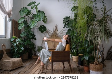 Overworked woman freelancer sitting on chair in cozy greenhouse, resting, stretching arms with closed eyes. Smiling tired woman in home garden taking break from online study. Plant lovers concept.