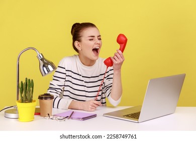 Overworked woman holding red retro phone and screaming with anger, nervous about deadline, lot of work, sitting at workplace with laptop. Indoor studio studio shot isolated on yellow background. - Shutterstock ID 2258749761