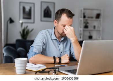 Overworked Young Entrepreneur In Tired From Work With A Laptop, Holds On To The Bridge Of The Nose His Eyes Closed. Stressed Guy Works Remotely At Home Feels Eyes Strain