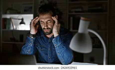 Overworked young hispanic man stressed from laptop work, grapples with office problem, dark night looms as pain from headache hurts, light from lamp reflected on his handsome beard - Powered by Shutterstock