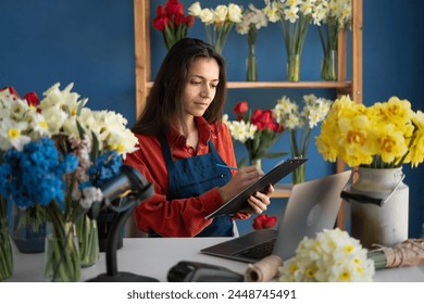 Own business concept. Happy Latin businesswoman in apron in small floral shop and writing down order in clipboard. Joyful female florist at work. Copy space - Powered by Shutterstock