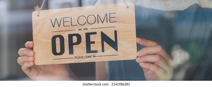 Owner business retail, asian young woman hand in turning sign board to open coffee shop, wearing face mask protect to pandemic of coronavirus, reopen store after close lockdown quarantine in covid-19. - Shutterstock ID 2141986281