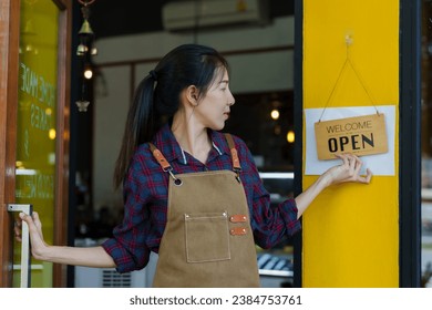 Owner of  coffee shop, small business, grabbed the door and turned and held up a welcome sign. Open shop in the morning And preparing to welcome customers, a beautiful Asian barista wears an apron. - Powered by Shutterstock