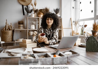 Owner of pottery workshop updates database of items that sold on website, sitting at table against background of shelves with products. Caucasian businesswoman engaged in filling social networks. - Powered by Shutterstock