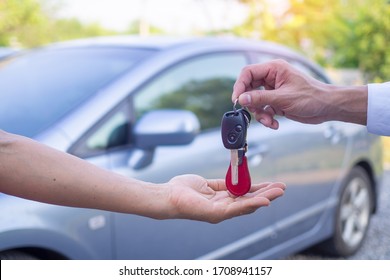 The Owner Sent The Car Keys To The Renter's Hands. There Is A Car Parked In The Back. Rent Or Buy Concept