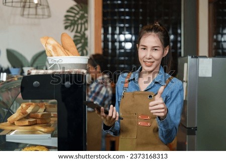 The owner of small cafe is beautiful Asian business woman. Standing behind the bakery phone in hand, gave thumbs up smile. There  baguette in the laundry basket. Retired mother making coffee the back.