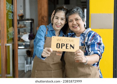 The owner of a small coffee shop, a retired woman, embraces and holds the head of her cute Asian daughter. Both held welcome and open signs. At the family coffee shop - Powered by Shutterstock