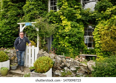 Owner Standing At The Front Gate With Garden Of Tubbs Mill House Bed And Breakfast Covered In Greenery At Mevagissey, Cornwall, England - June 11, 2019